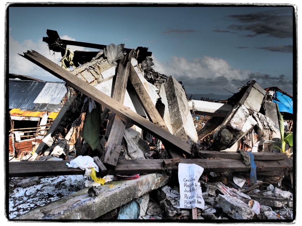 A list of names on a sign sits in front of the remains of a house